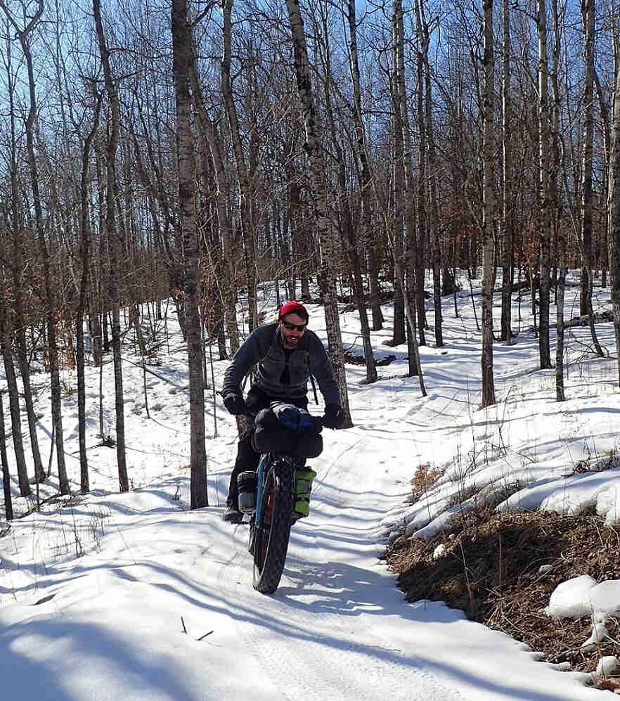 Front view of a cyclist riding a turquoise Surly Wednesday fat bike with gear, down a snow covered trail in the woods
