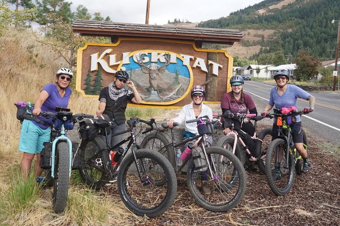 Group of 5 cyclists standing with their bikes with gear in front of a Kligkitat sign on  the shoulder a mountain road