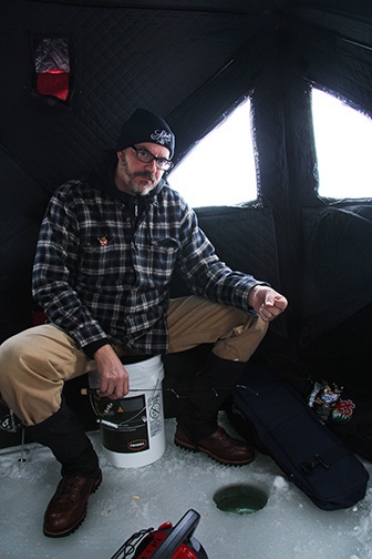 Front view of a person sitting on a bucket, over a hole in the ice, inside of an ice fishing shelter