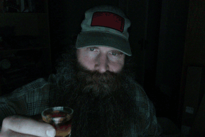 Front view of a person with a large beard, wearing a baseball cap, taking a shot of liquor in the dark