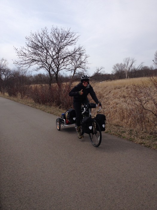 Front, right side view of a cyclist, riding a Surly bike with trailer down a paved trail with a grass field behind