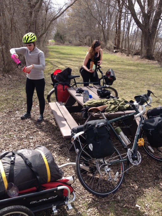 Two cyclists standing around a picnic table with their Surly bikes and gear, on a grass clearing in the bare woods