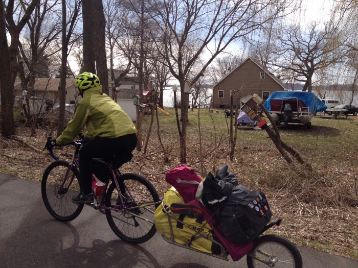 Left side view of a cyclist, riding a Surly bike with trailer on a paved trail, past the back of a house on a lake