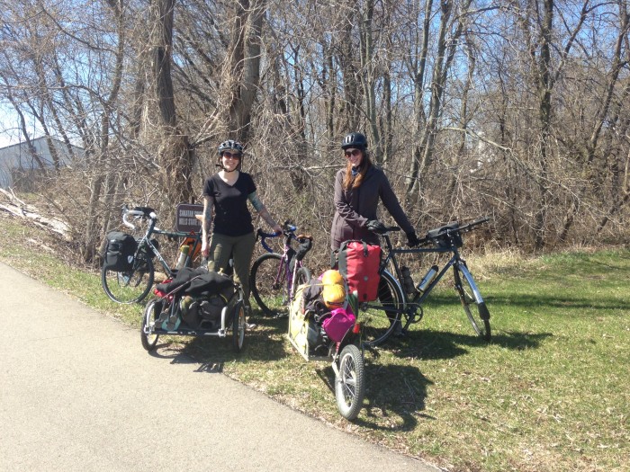 Two cyclists posing with their Surly bikes and trailers, on the side a paved trail, on grass with bare trees behind them