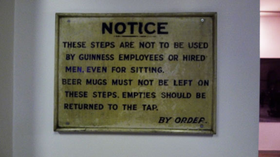 A workplace notice sign, with black text and yellow background, posted on an interior wall
