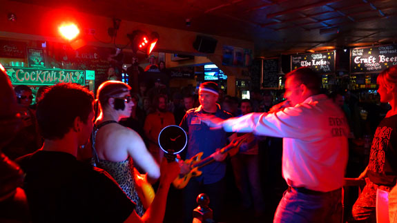 Left side view of a person singing into a microphone, in a dimly lit bar with people standing around watching