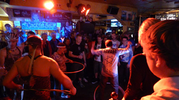 People in a dimly lit bar, competing in a hula hoop contest, with others standing around watching