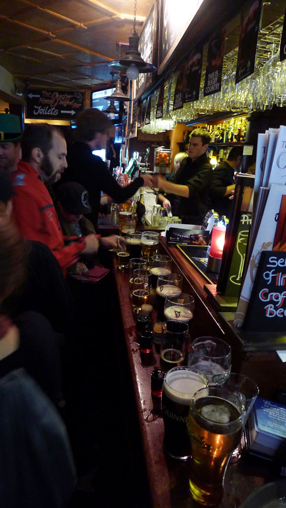 Side view of glasses of beer lined up on a counter top in a bar with people standing around