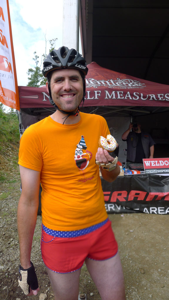 Front view of person wearing a bike helmet, red shorts and an orange t-shirt with a clown graphic, eating a donut