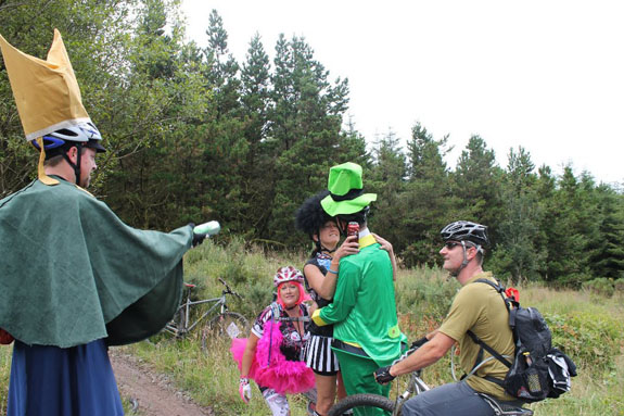 Left side view of a cyclist sitting on a bike, next to people in costumes, on a dirt trail with trees in the background