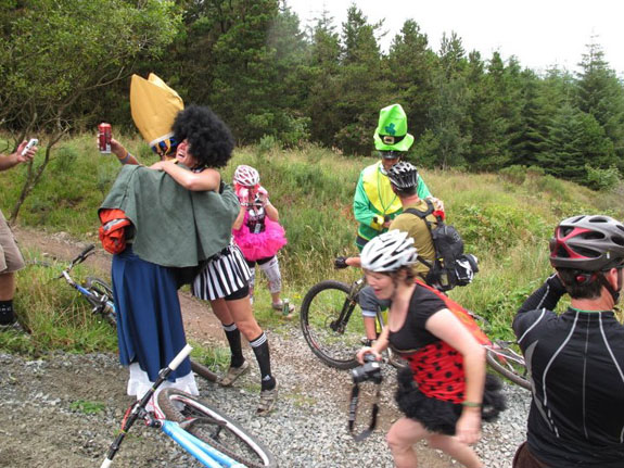 A group of people in costumes, and cyclists riding bikes, on a dirt trail with trees in the background
