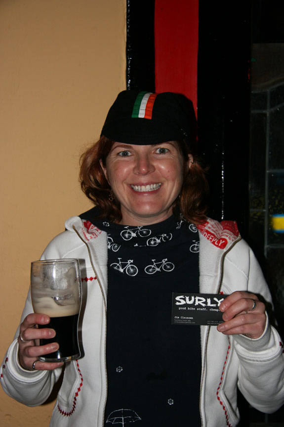 Front, waist up view of a smiling person, holding a pint of Guinness beer and a Surly business card in their hands