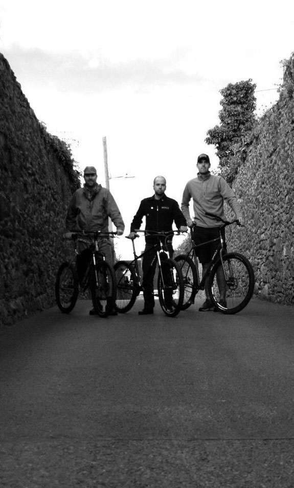 Front, right side view of 3 Surly bikes, with 3 cyclists standing with them, on a road with stone walls on the sides