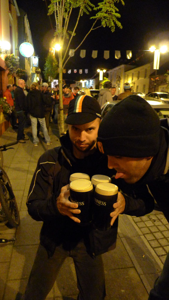 Front view of a person holding 4 glasses of Guinness, with other people standing around them, on a sidewalk at night