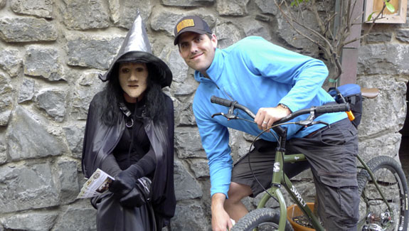 Front left side view of Surly Ogre bike with a cyclist on it, next to a person in a witch costume, against a stone wall