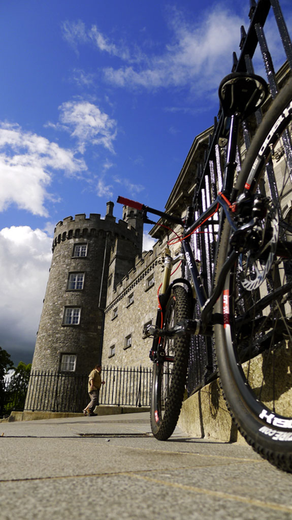 Ground up, rear view of a black Surly bike, leaning against a rail on a paved parking lot, next to a castle