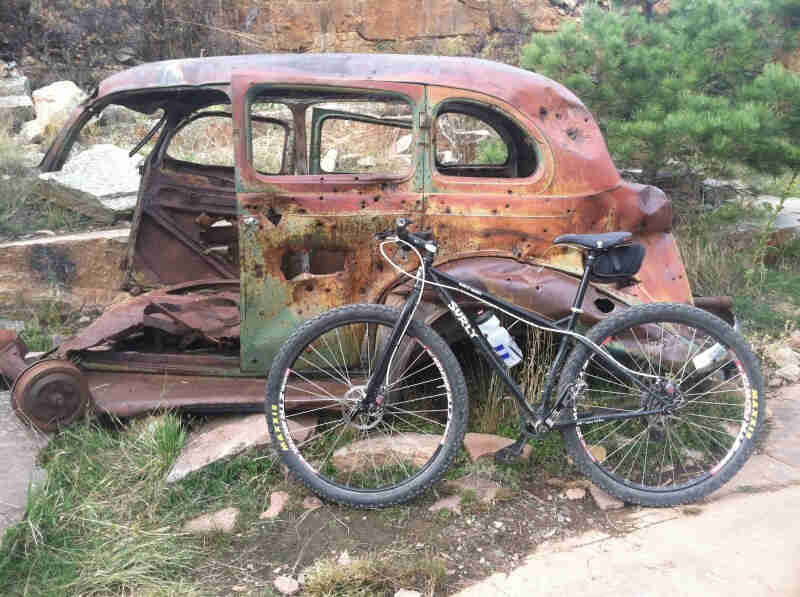 Left side view of a black Surly Karate Monkey bike, parked on the left side of an old car body with bullet holes