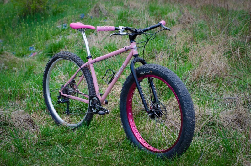 Right side view of a pink Surly 1x1 bike with a fat wheel on front, parked in a grass field
