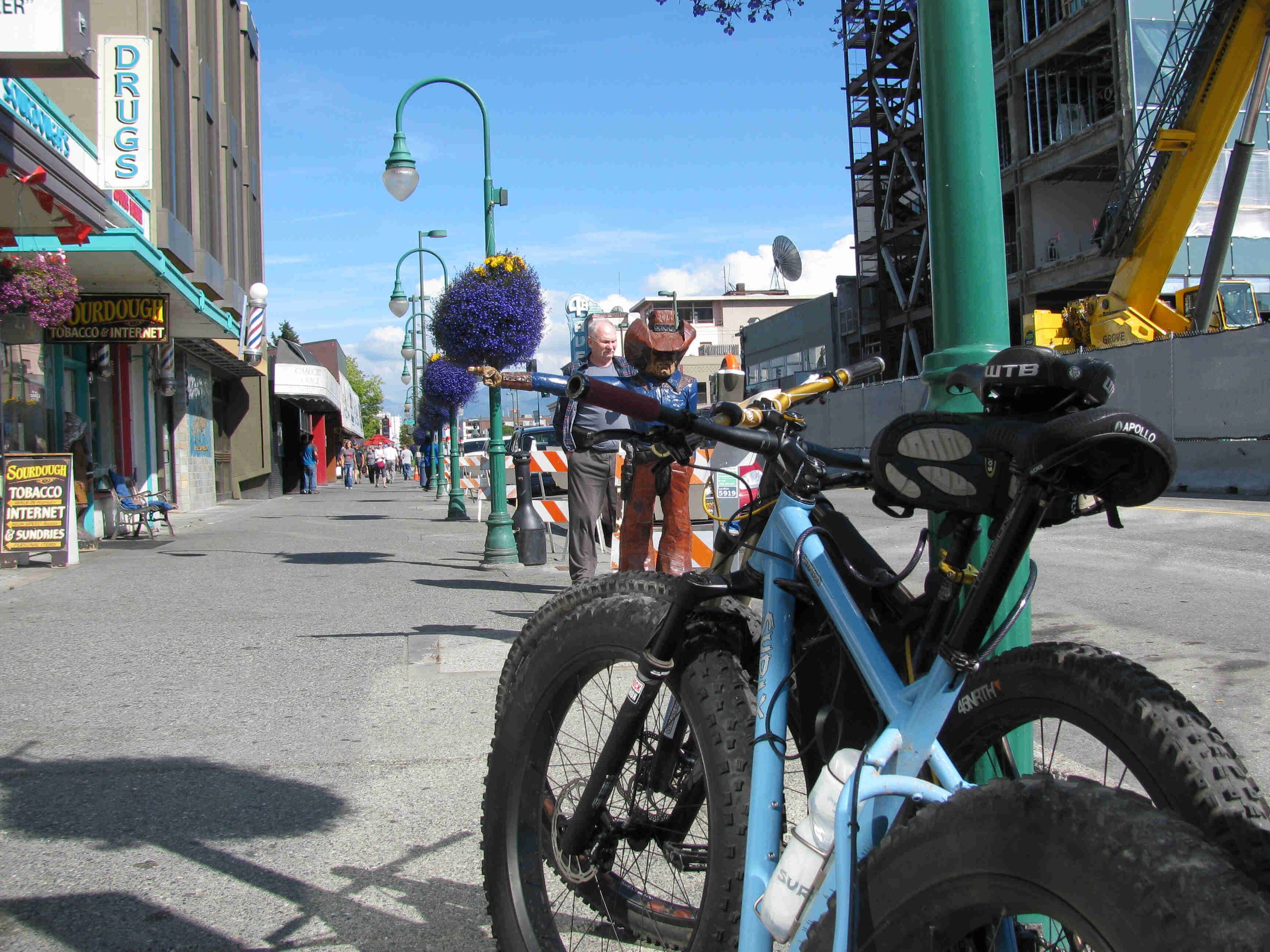 Rear, left side view of 2 Surly fat bikes, parked side by side, against a light pole on a sidewalk with city shops
