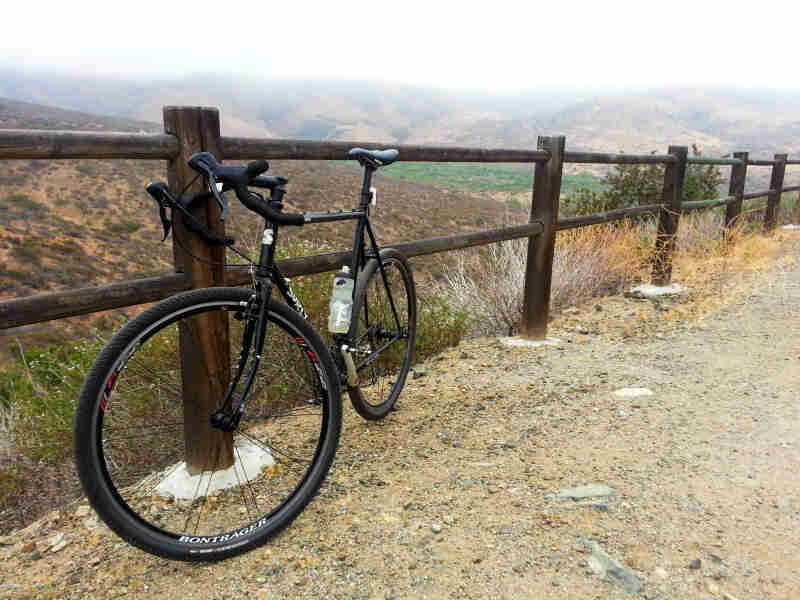 Front, left side view of a black Surly bike, leaning on a wood post fence, with fog covered mountains in the background