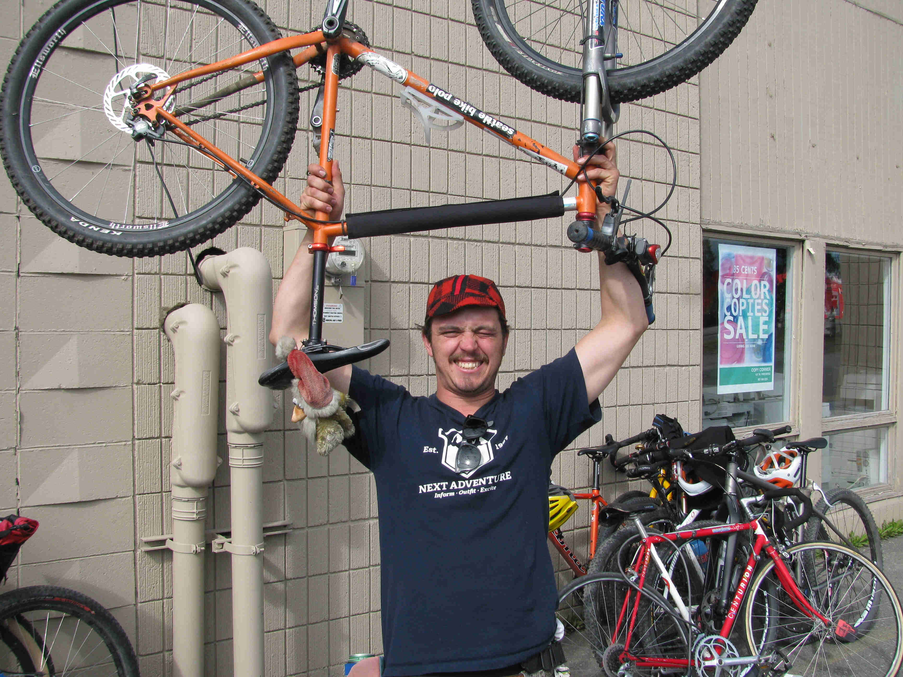 Front view of a person, holding an upside down, bike over their head, with bikes stacked against a wall behind them