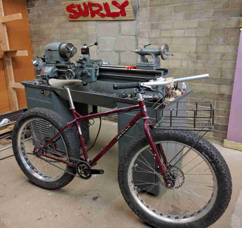 Right side view of a Surly Pugsley bike, maroon, parked on workshop floor, leaning against a tooling machine  