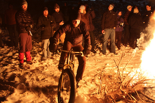Front view of a cyclist riding a Surly Pugsley fat bike on snow, around a campfire at night, with people behind