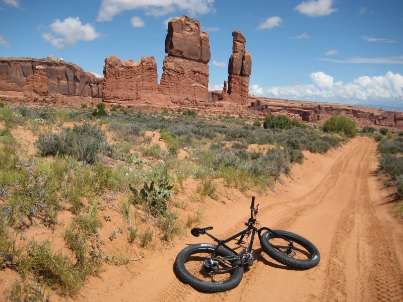 Rear view of a black Surly Pugsley fat bike, laying down on a sandy road, facing down a desert field with buttes ahead