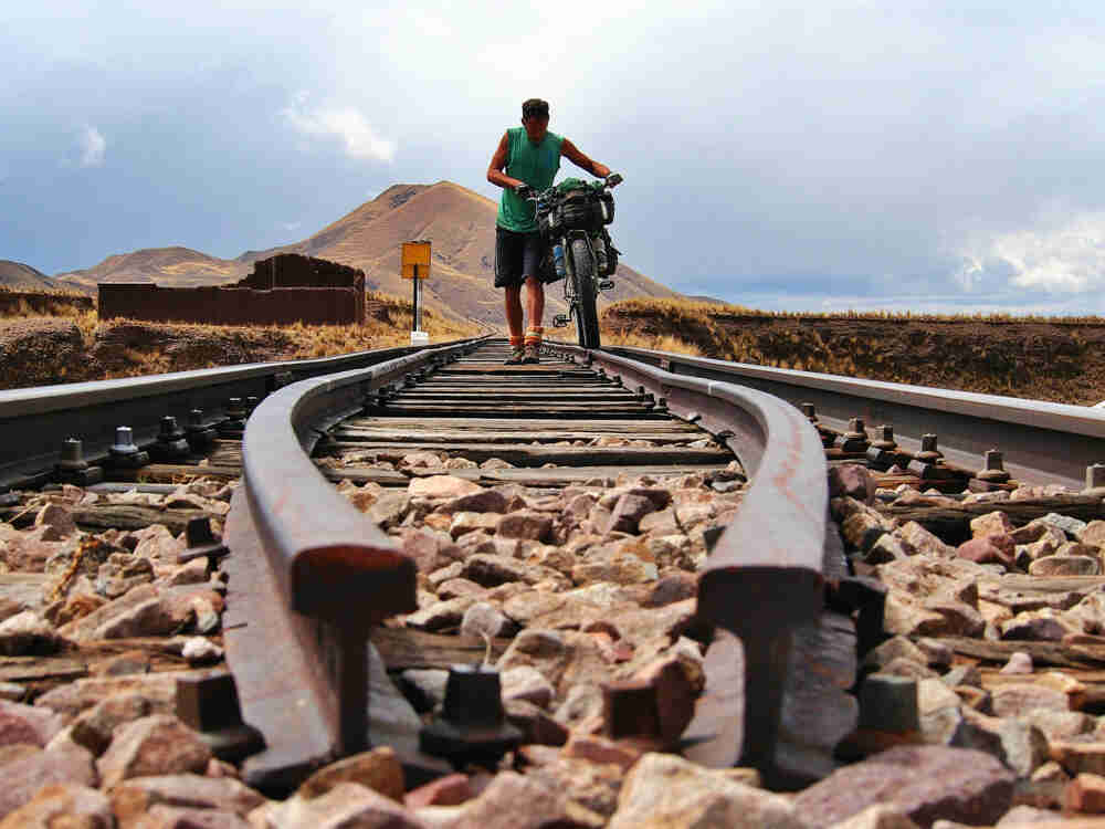 Front view of a cyclist, walking a Surly Pugsley bike with gear, on railroad tracks with hills in the background