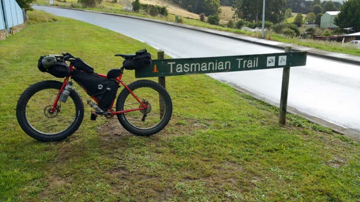 Left side view of a red Surly Pugsley fat bike, loaded with gear, parked on grass, leaning on a Tasmanian Trail sign