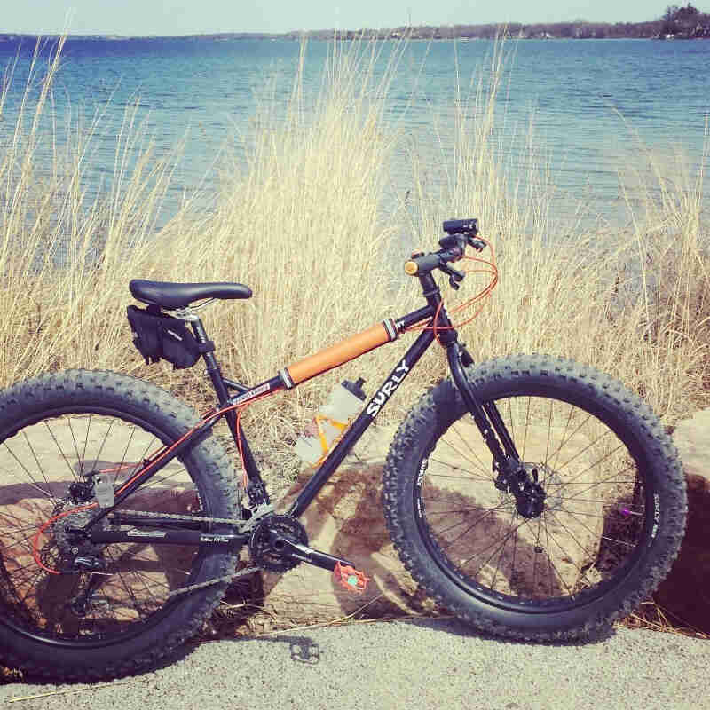 Right side view of a black Surly Pugsley fat bike, parked against rocks in front of tall grass with a lake behind