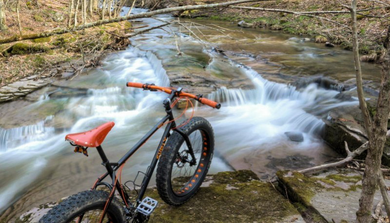 Downward right side view of a Surly Pugsley fat bike, parked on the bank of a rocky river, in a fall forest