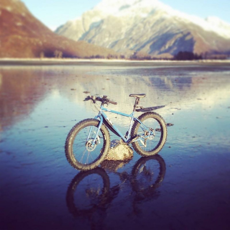 Left side view of a blue Surly Pugsley fat bike, standing over a rock on a frozen lake with mountains in the background
