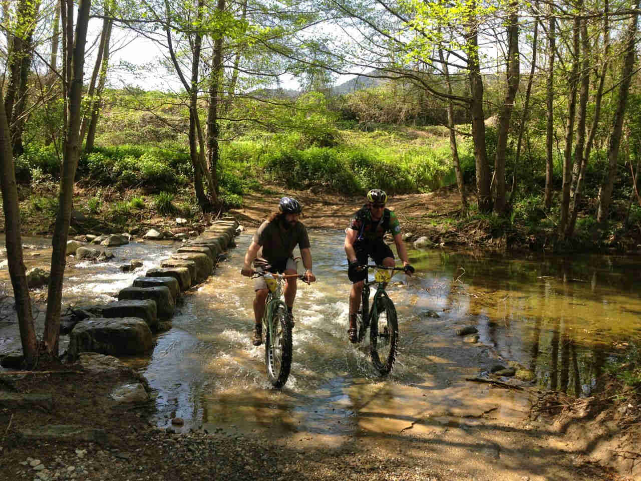 Front view of 2 cyclists, riding their bikes across a shallow river,, in the woods