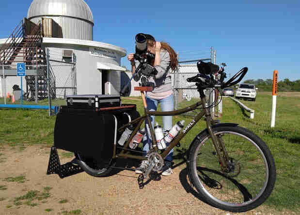 Right side view of an olive Surly Big Dummy bike, with a person using the seat post to mount a telescope