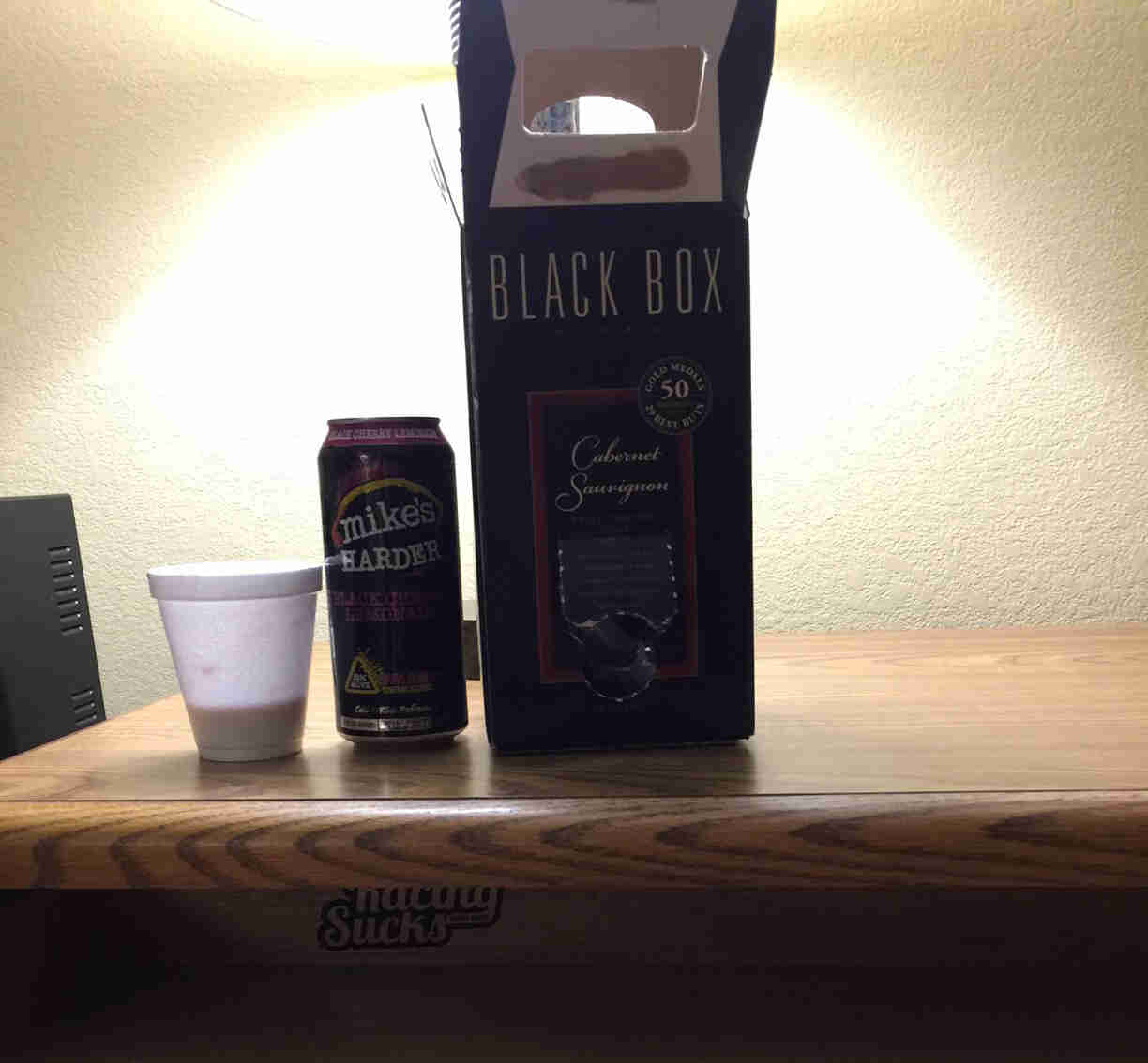 A styrofoam cup, a beverage can and a box of wine on a table top, with wall in the background
