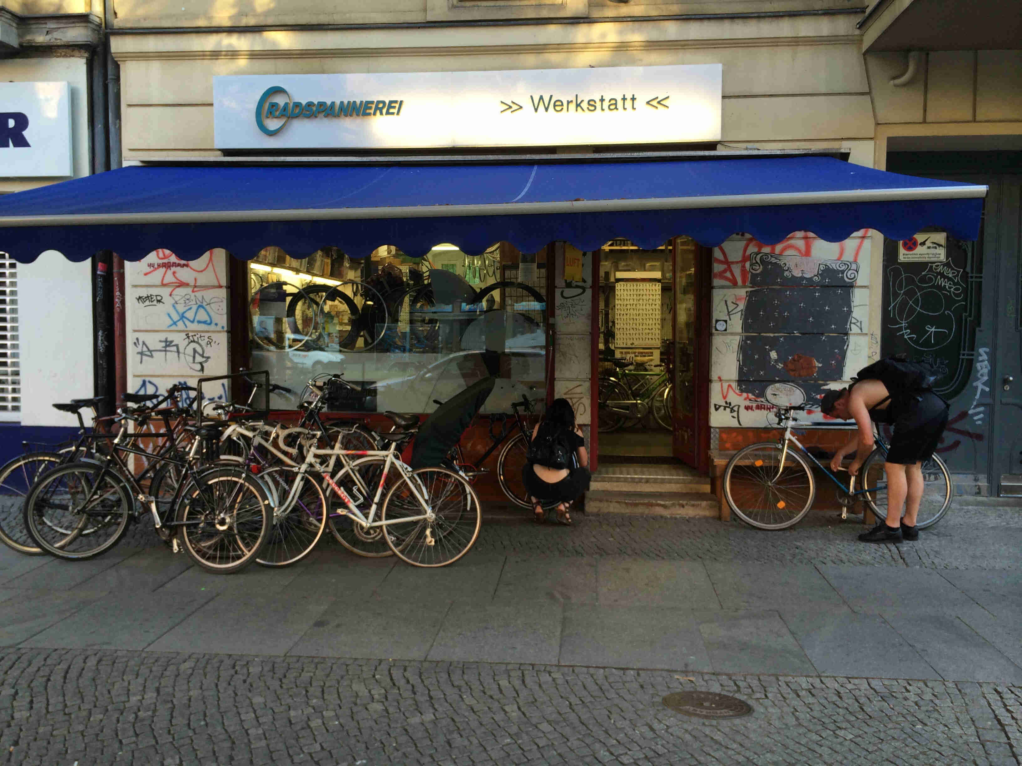 Front view of a group of bikes, parked side by side on a brick sidewalk, in front of the Radspannerei bike shop