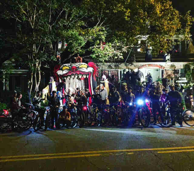 Front view of a group of cyclists, lined up side by side, posing for a picture at night, in front of a haunted house