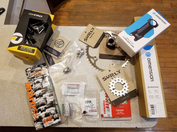 Downward view of a table on a wood floor covered with Surly and other brands of bike parts and boxes