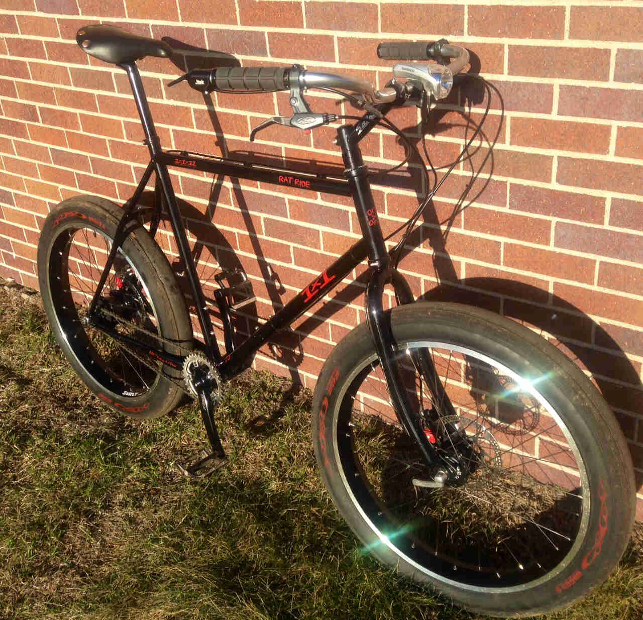 Right side view of a black Surly 1x1 Rat Race bike, on grass, leaning against a brick wall