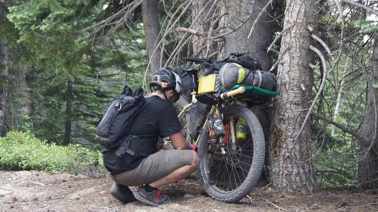 Front right side view of a cyclist squatting down next to a fully loaded bike against a tree in the forest