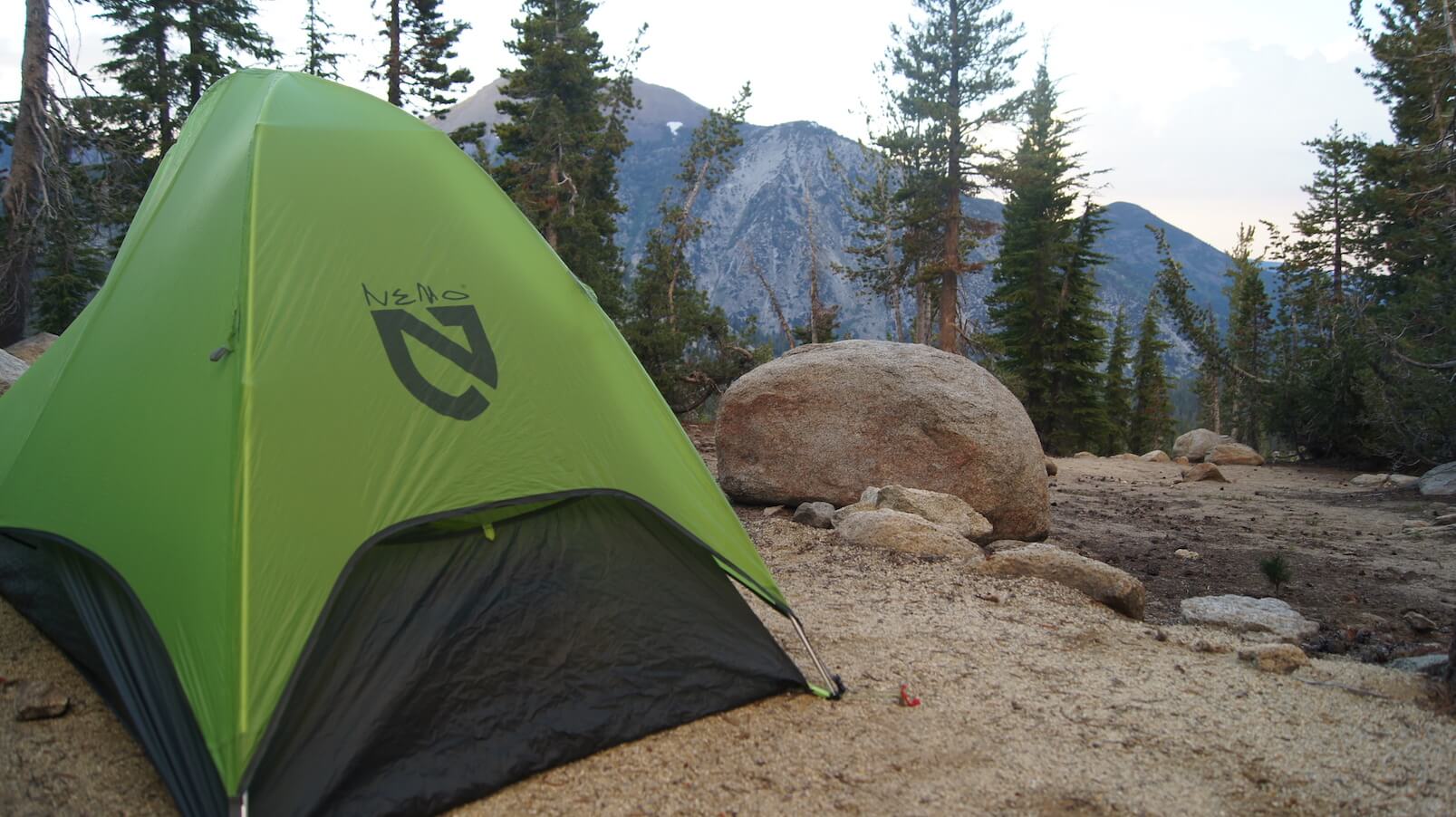 A green tent on a sandy gravel plot with a boulder behind, with pine trees and mountains in the background