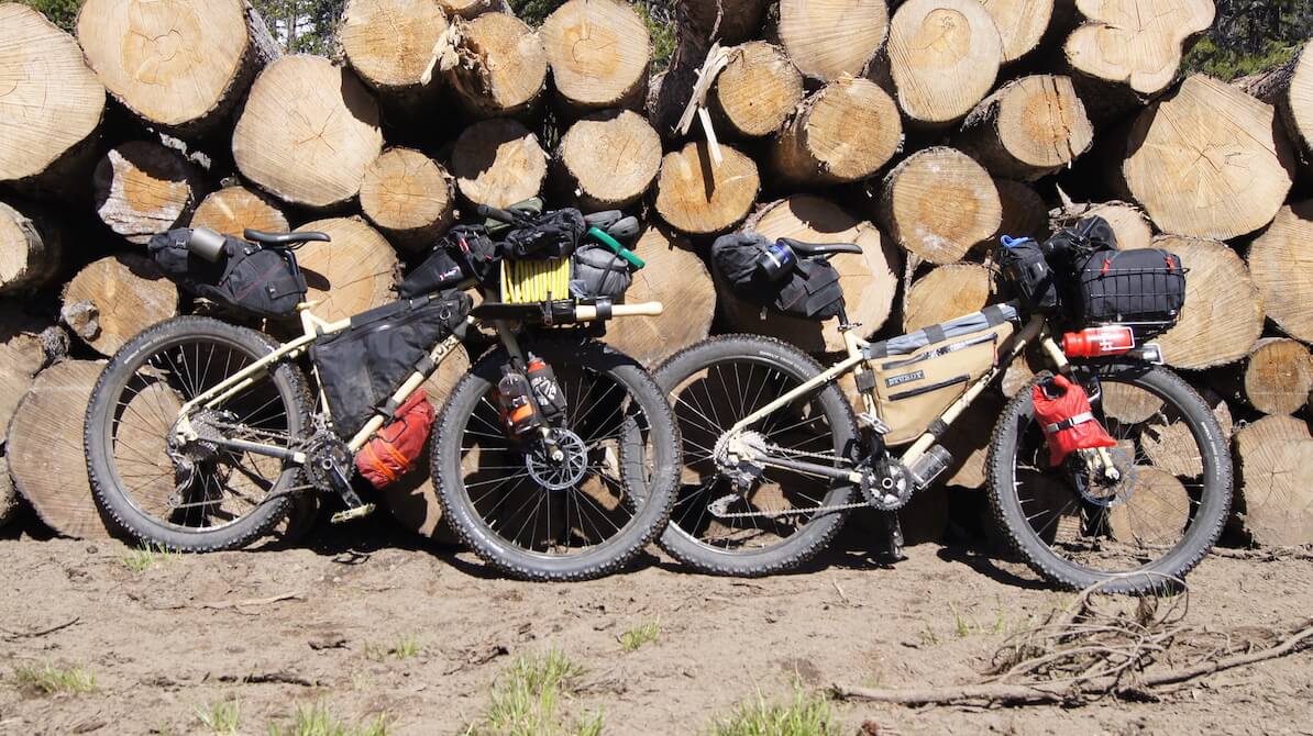Two Surly bikes, loaded with gear, on sand, leaning on the end of a stack of logs 