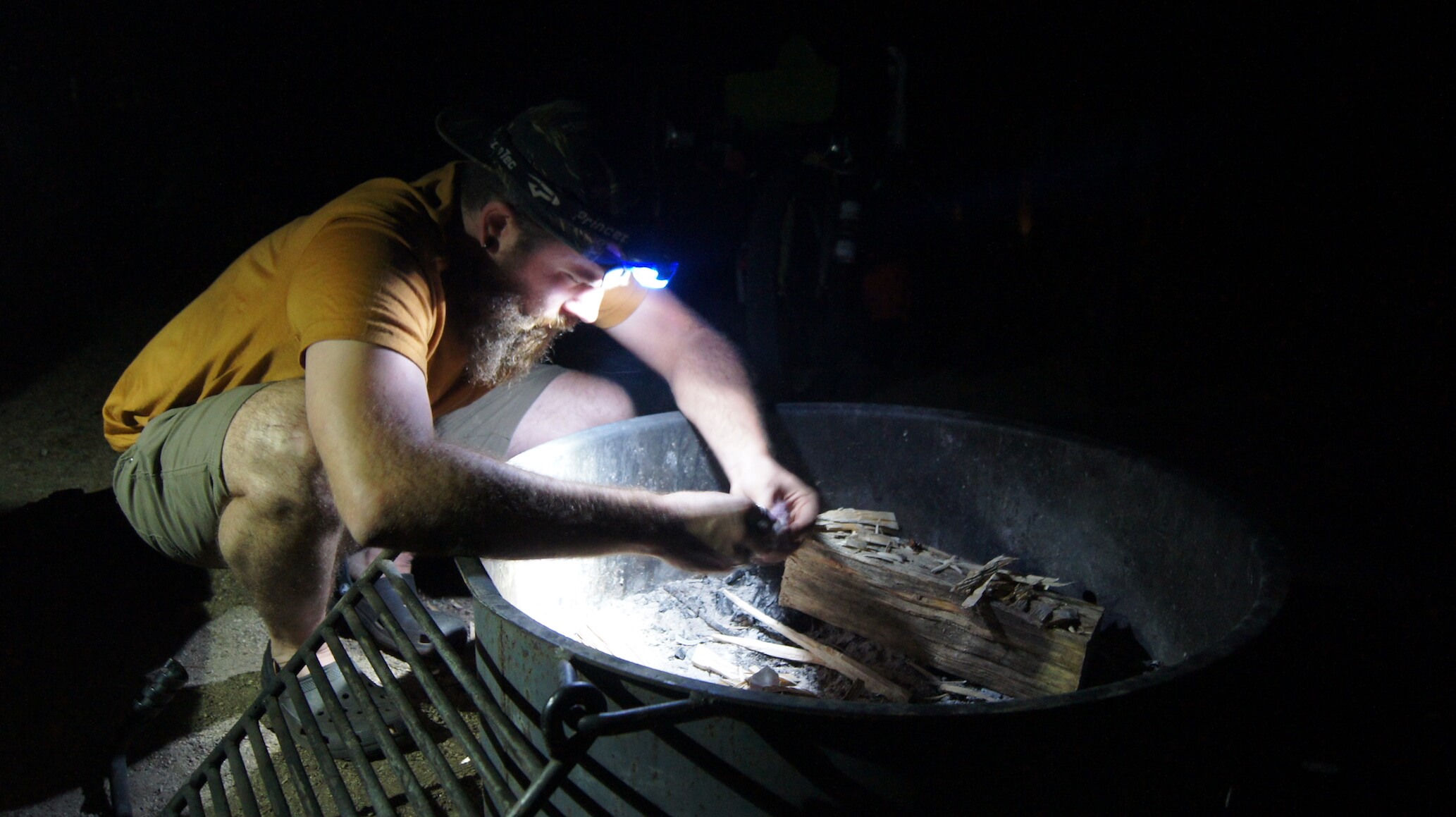 A person wearing a headlamp, squats over a campfire ring at night