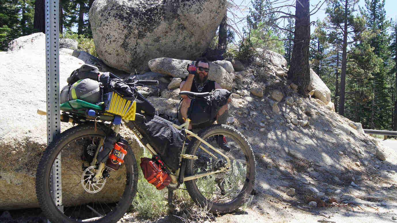 Left side view of a Surly bike in front of a cyclist sitting on a rock, with a boulder and pine trees in the background