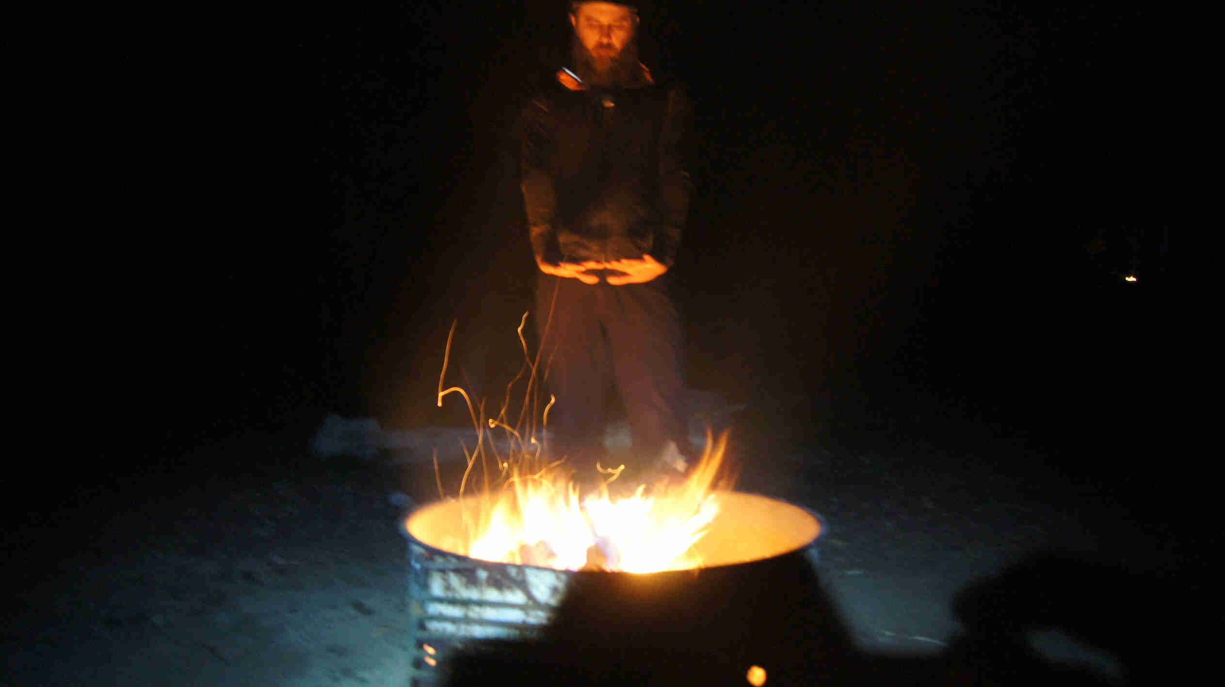 A person warming their hands above a fire in a burn barrel, at night