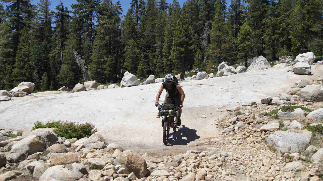 A cyclist rides a bike loaded with gear, down a large flat rock, with pine trees in the background