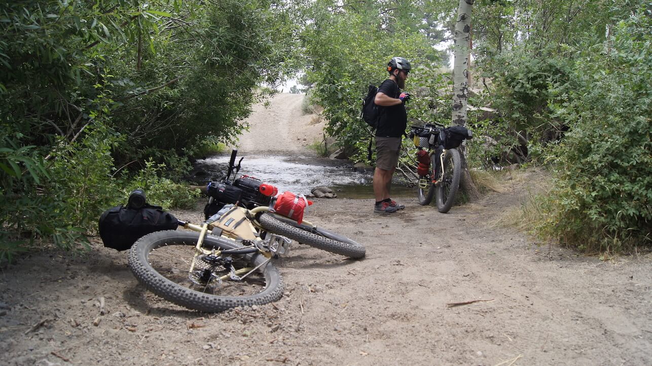 A cyclist standing on the left side of a bike in front of a river in the woods, with a bike leaving in sand behind