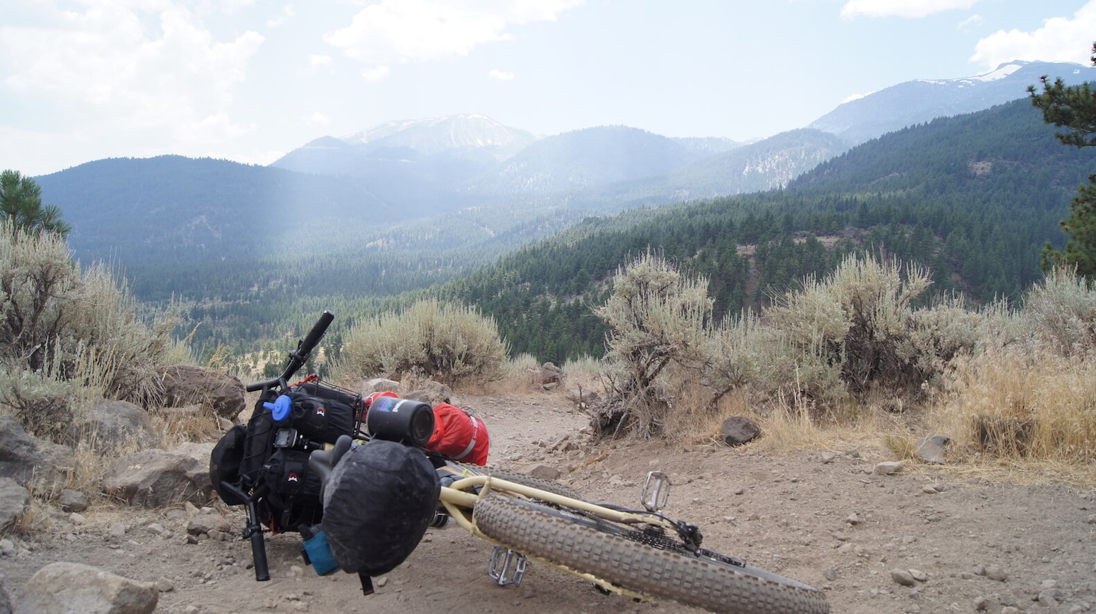 Rear view of a bike, loaded with gear, laying on its side on a gravel rocky trail, with foggy mountains ahead