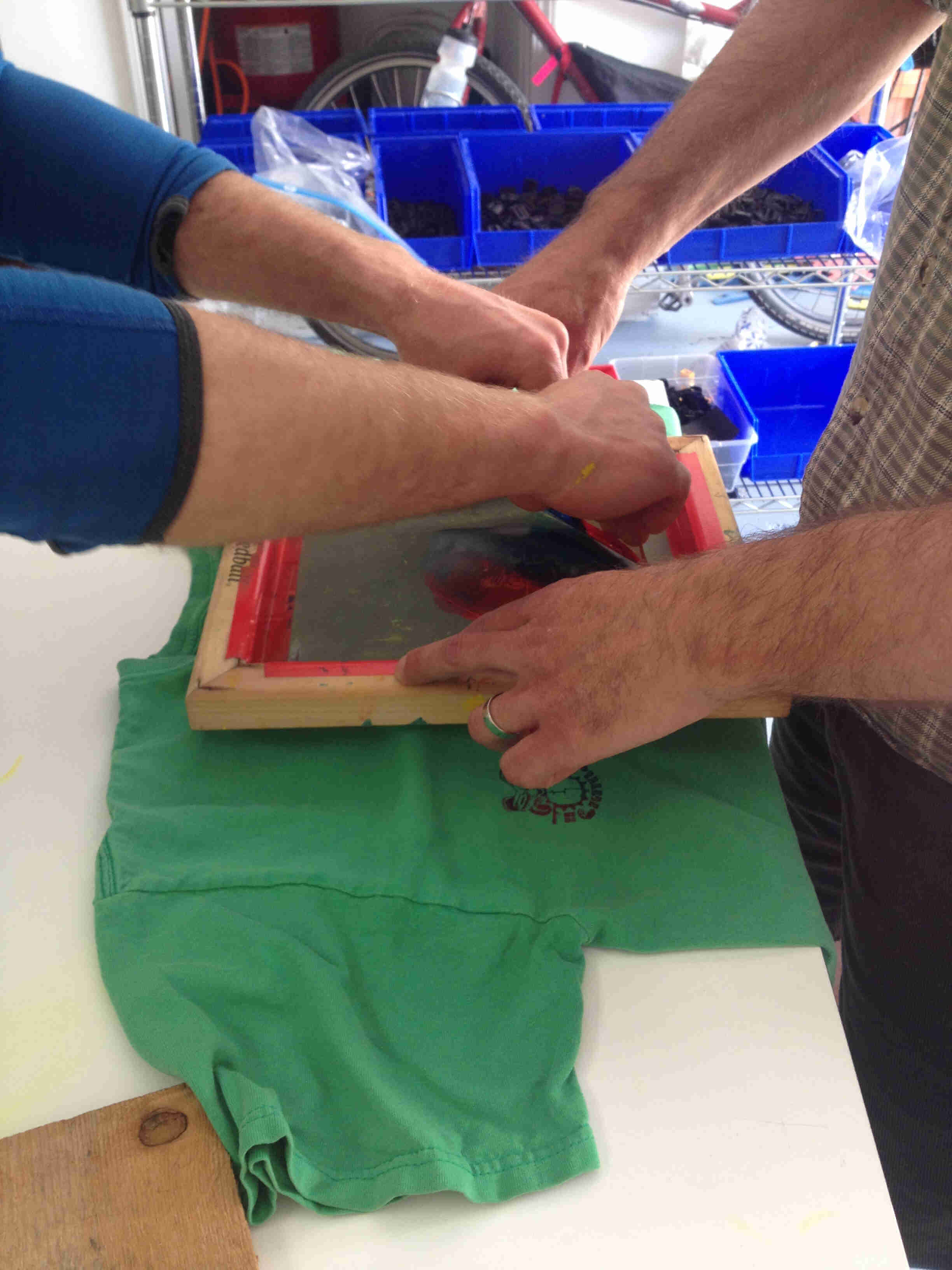 Side view of 2 sets of hands, from people standing across from each other, silk-screening a green t-shirt together